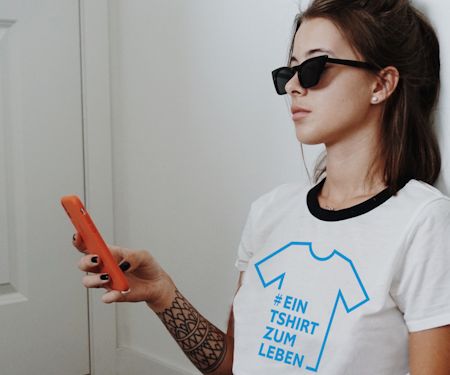 Frau mit Sonnenbrille hält Handy und trägt weißes T-Shirt mit blauer Aufschrift Ein T-Shirt zum Leben